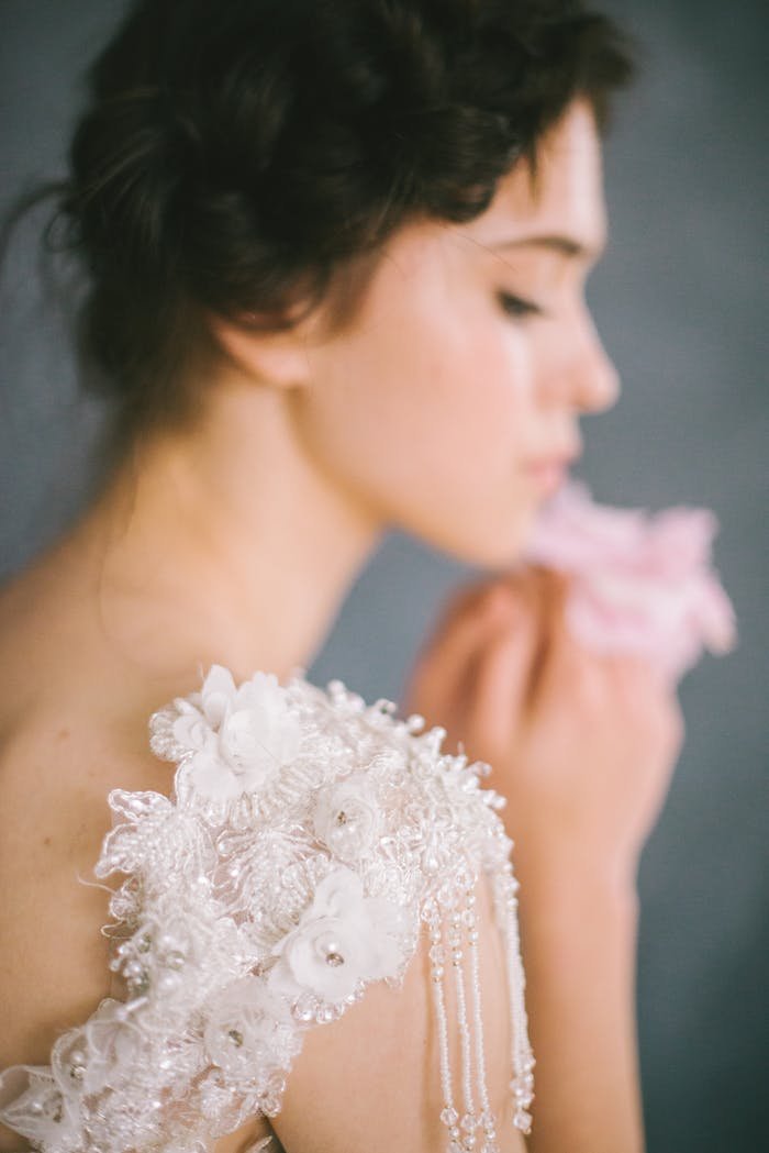 About Soft-focus portrait of an elegant bride holding a pink flower, wearing embroidered lace.